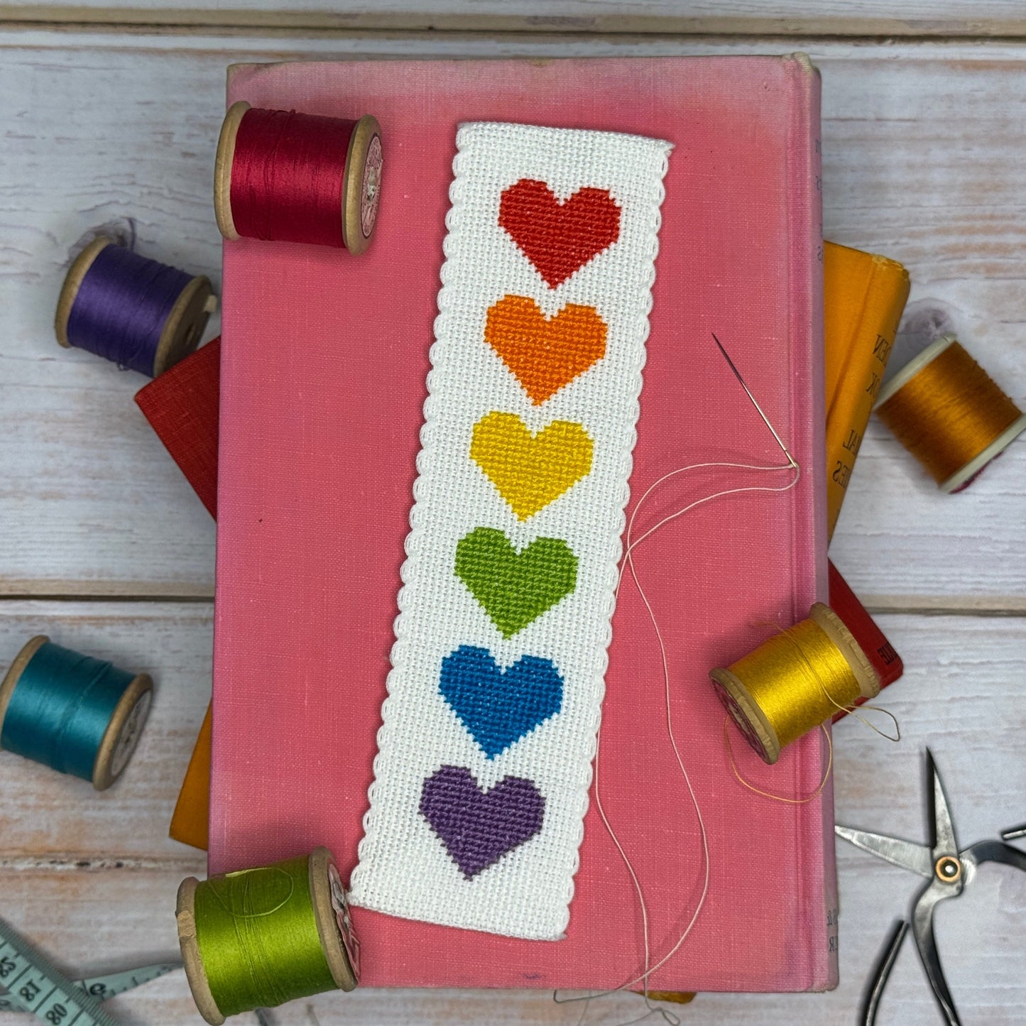 Embroidered bookmark with colorful hearts on a pink book surrounded by spools of thread and scissors.