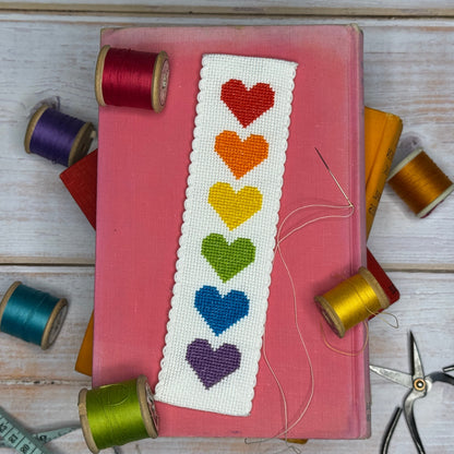 Embroidered bookmark with colorful hearts on a pink book surrounded by spools of thread and scissors.
